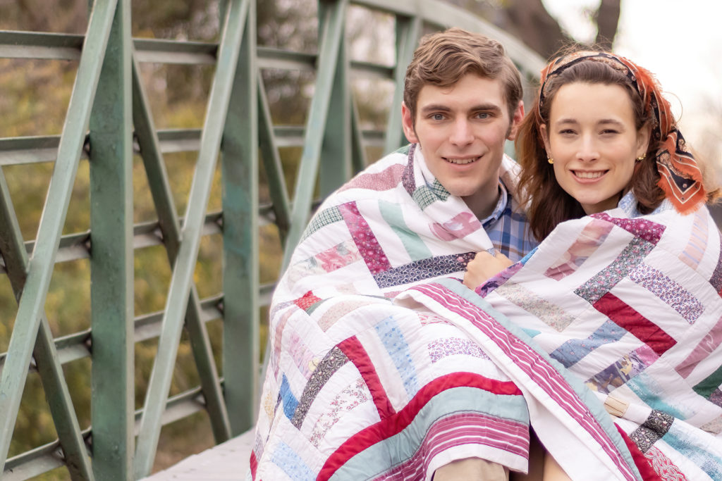A couple takes anniversary pictures on a bridge sitting down. They have a quilt wrapped around them.